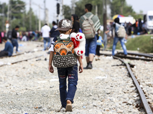 A child wearing a cap walks with a backpack and a stuffed animal at the US-Mexico border