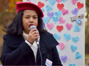 Ronnate speaks into a microphone at an outdoor event. She wears a coat and a red hat. Behind her is a board with heart-shaped sticky notes with writing on them. 