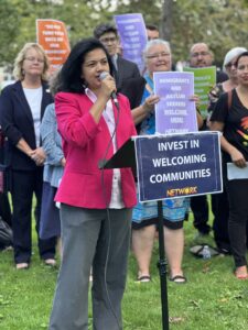 NETWORK Government Relations Director Ronnate Asirwatham, a woman in a pink jacket, holds a microphone and speaks from behind a podium with a sign, "Invest in Welcoming Communities." Many other advocates with similar signs stand behind her. 